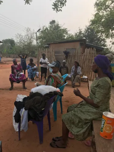 A woman addresses a gathering of women seated outdoors, with folded donated clothing on chairs in the foreground.