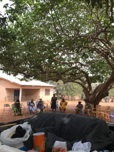 A meeting of women under a broad tree outside a house, viewed over a pickup truck carrying supplies.