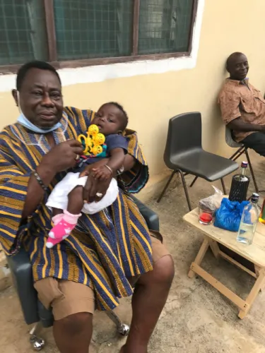 An older man in a striped kente smock holds a baby on his lap while seated outdoors.
