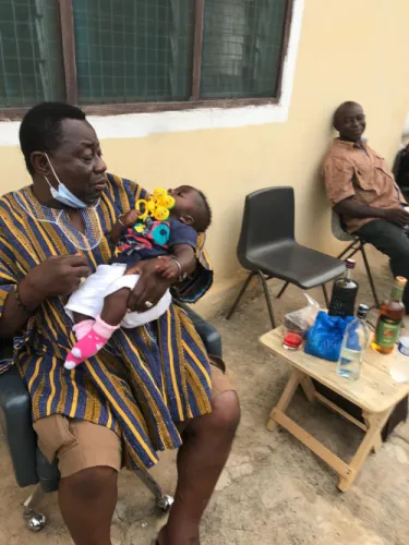 A man in traditional dress cradles an infant, seated outside a building with another man nearby.