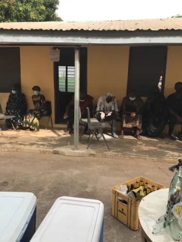 Community members seated along the veranda of a yellow building at a gathering, with crates of drinks in the foreground.