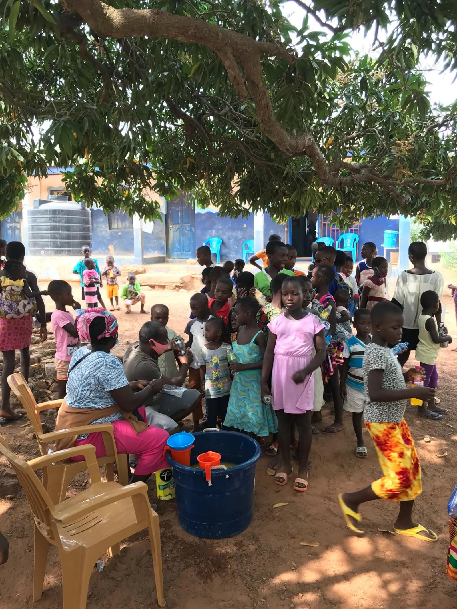 Children queuing under a mango tree outside a blue-painted school while volunteers hand out bottled drinks from a large tub.