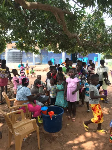 Children queuing under a mango tree outside a blue-painted school while volunteers hand out bottled drinks from a large tub.