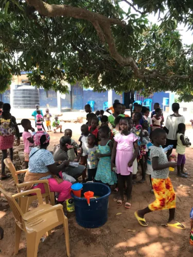 Children gathered under a mango tree at a drinks distribution outside a blue-painted school building.