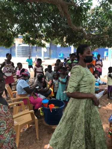 A volunteer in a green dress carries supplies past a group of children waiting under a tree for drinks to be handed out.
