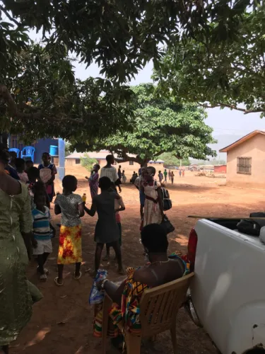 Children and adults gathered under mango trees at the edge of a village, with a pickup truck parked alongside.