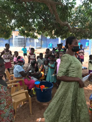 Volunteers distribute bottled drinks to children from a tub under a mango tree outside a school.