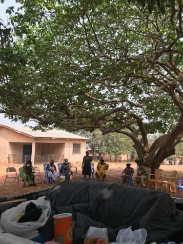 Women seated in a circle of chairs under a large tree outside a house, seen past the loaded bed of a pickup truck.