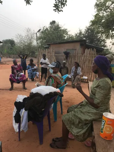 A woman in a green dress speaks to a seated group of women outdoors, beside chairs piled with donated clothing.