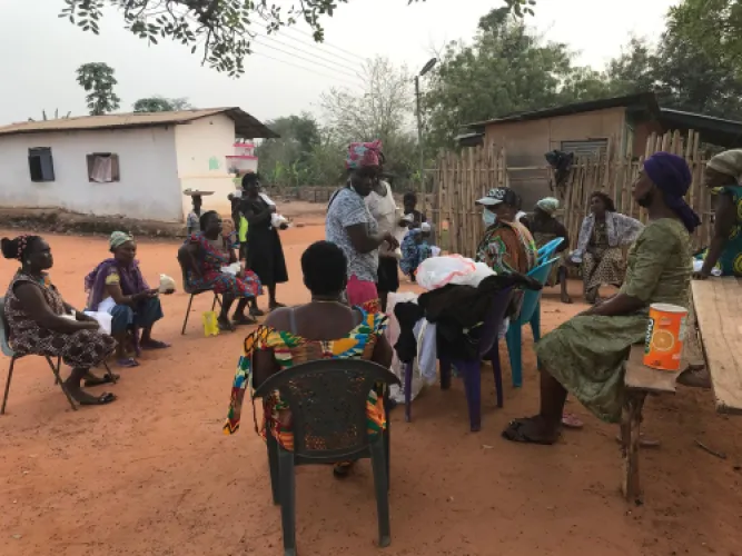 Women seated in a wide circle on bare ground, with donated clothing and supplies stacked on chairs between them.